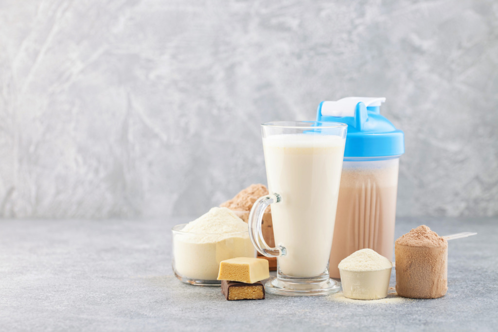 Sports nutrition, fitness diet and food concept - glass, protein shake bottle, powder and bars on grey background