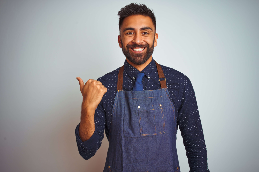 Young indian employee man wearing apron uniform standing over isolated white background smiling with happy face looking and pointing to the side with thumb up.